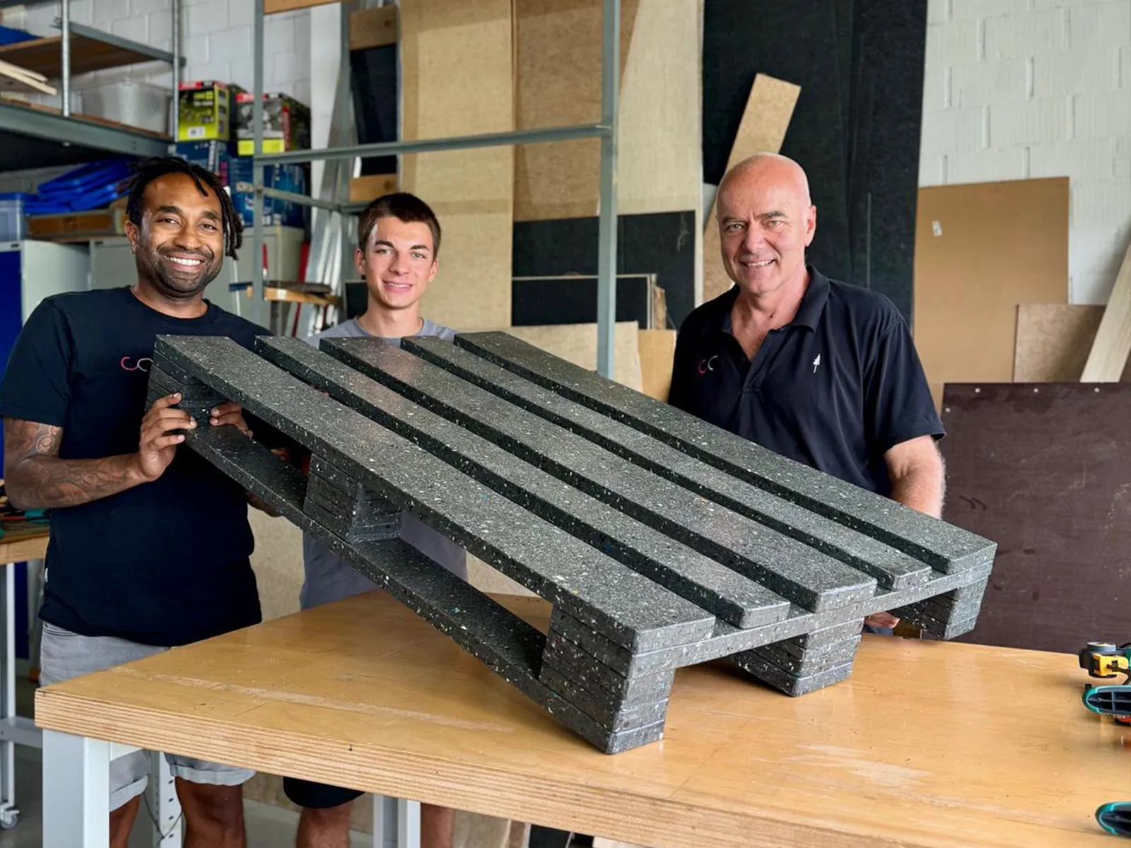 Three people presenting an assembled dark composite pallet prototype on a workshop table